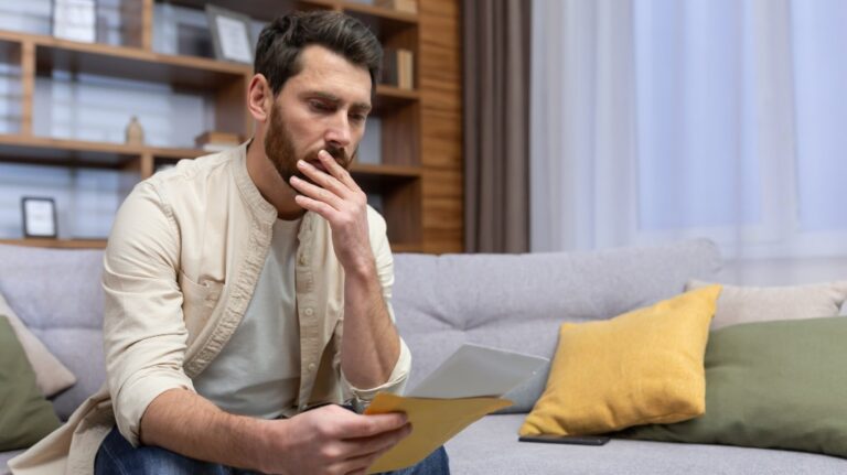 Shocked young man sitting on the sofa at home, holding a letter in his hands