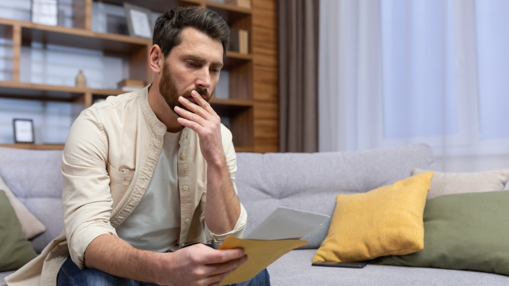 Shocked young man sitting on the sofa at home, holding a letter in his hands